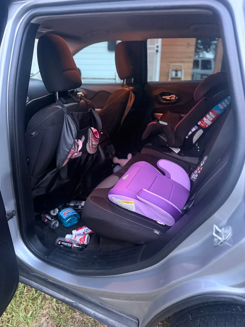 Rear seat of a silver Jeep Cherokee before detailing, with two child car seats, a seat back organizer and drink bottles on the floor
